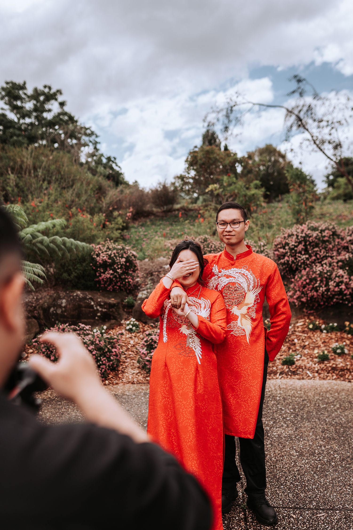 A photoshoot of a couple wearing matching red traditional Chinese outfits with embroidered details in a garden setting.