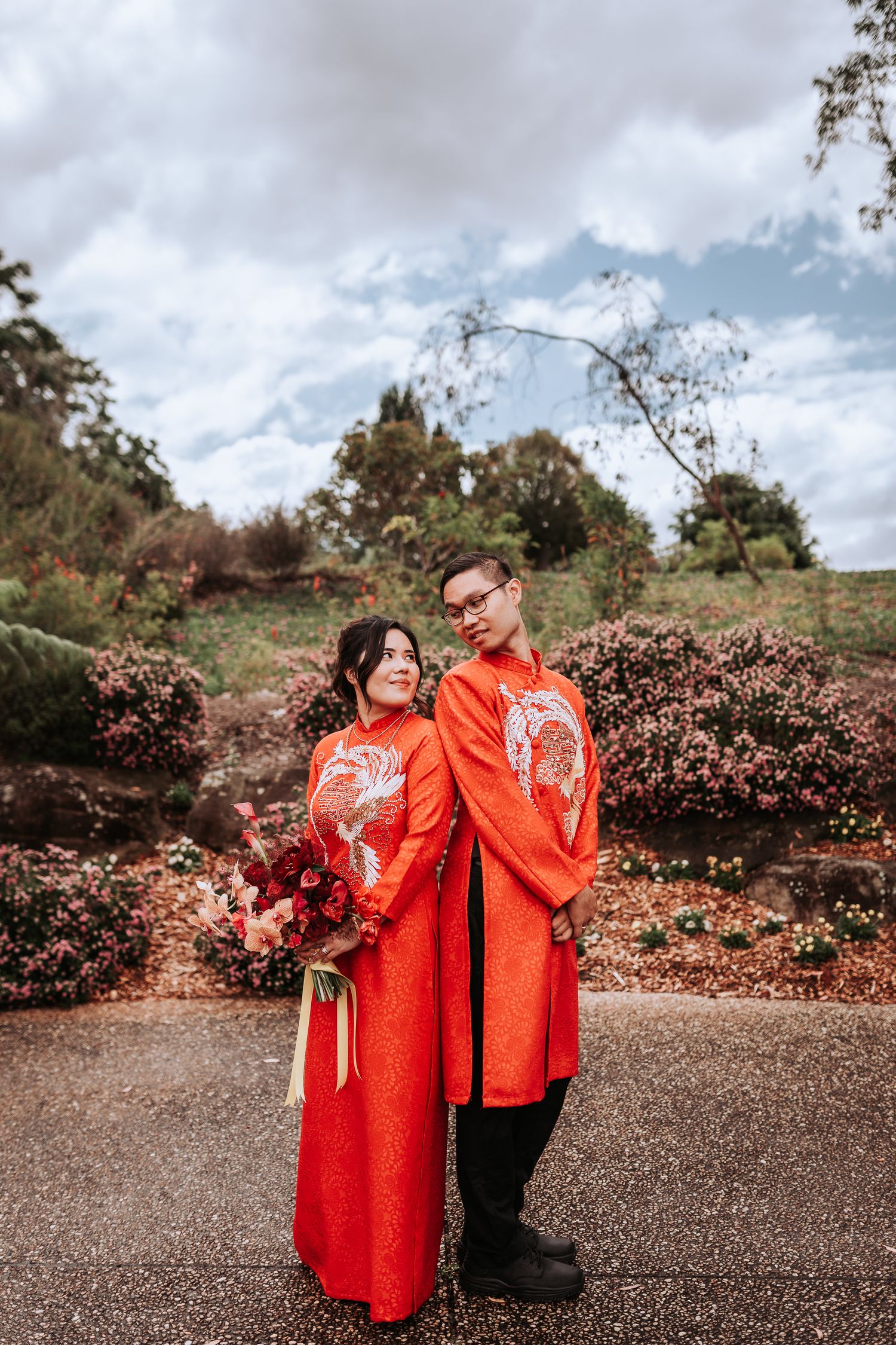 Couple in matching bright red traditional Asian wedding attire stand together in a garden with cloudy sky background.