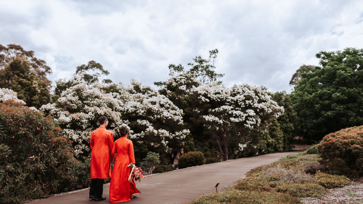 Two people in vibrant red traditional outfits walking along garden path with blooming white flowers.