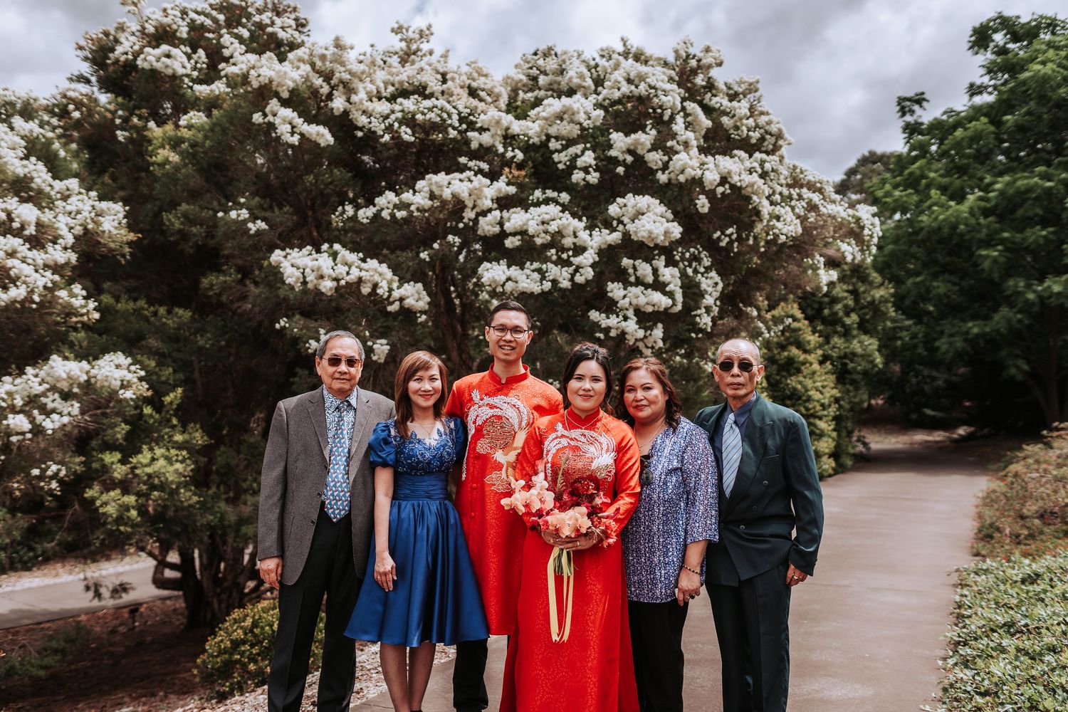 A wedding party poses together in front of blooming white flowers with the couple in red traditional attire.