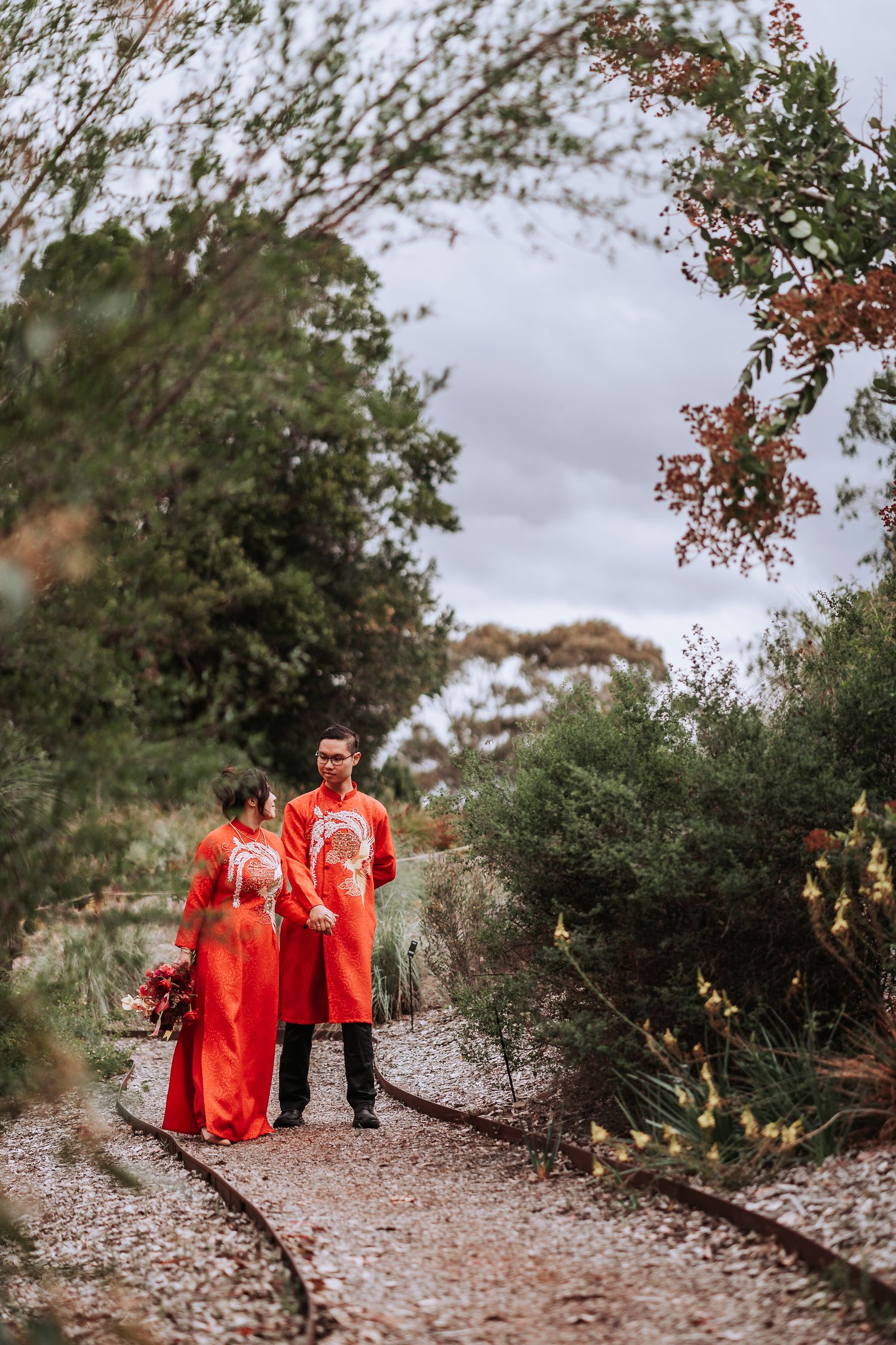 A couple in matching red attire walks along a rustic garden path surrounded by greenery.