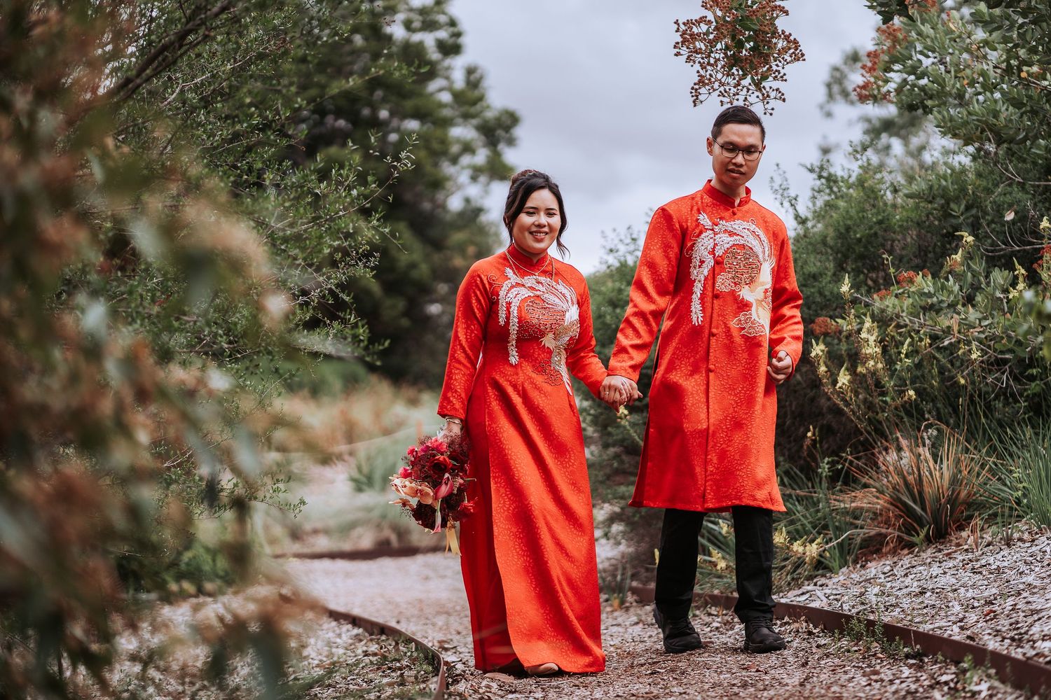 A couple in traditional red Vietnamese wedding ao dai attire pose together on a garden path.