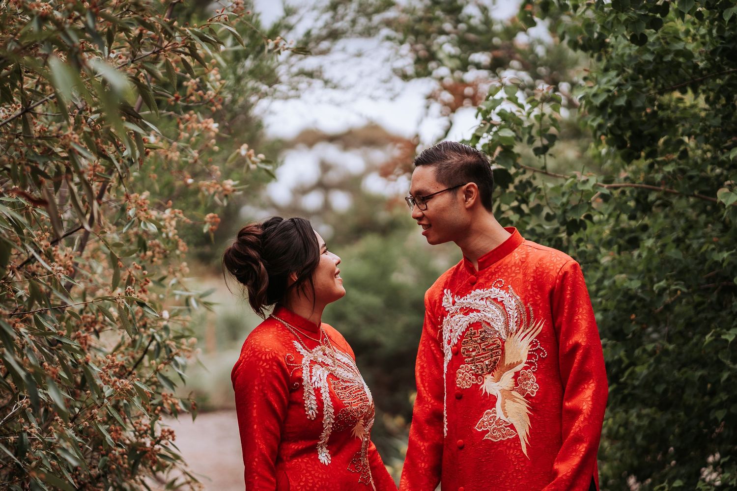 Couple in matching red Chinese traditional wedding outfits with phoenix embroidery stand together in garden setting.