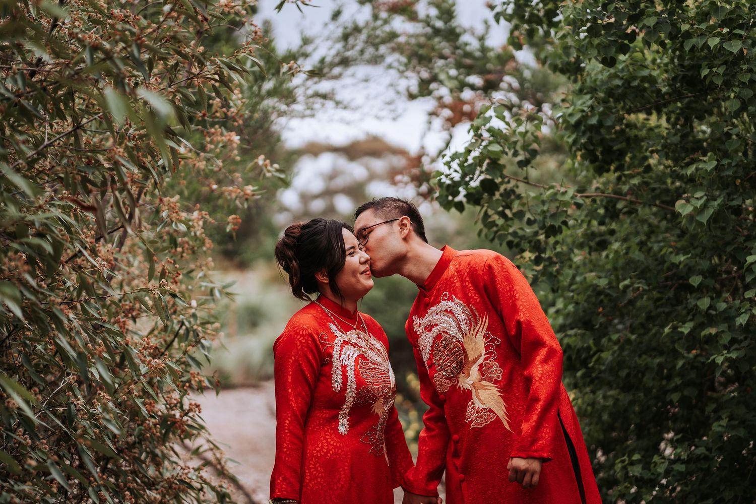 Newlyweds in matching red Chinese traditional attire share intimate moment in garden setting.