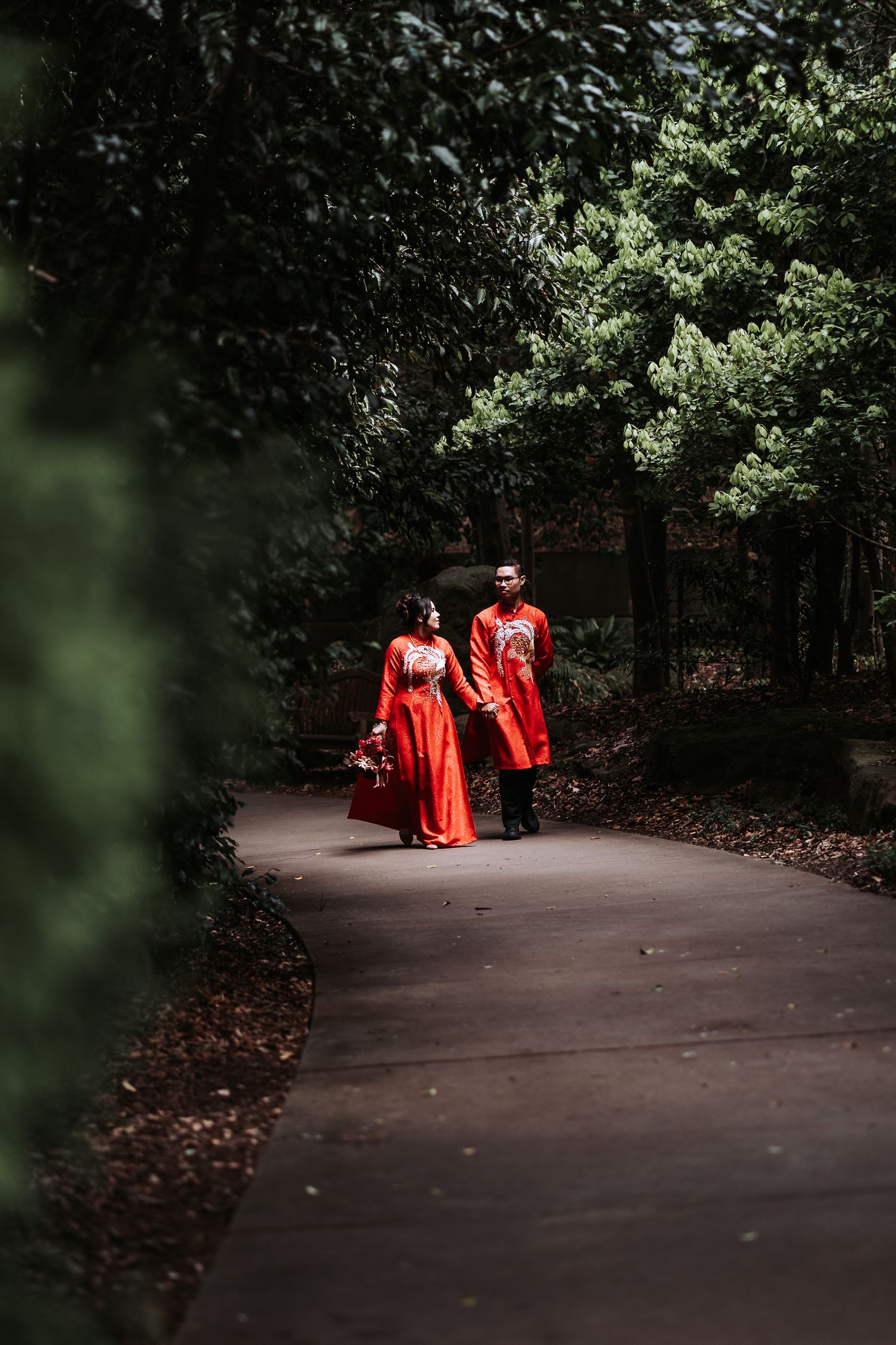 Two figures in flowing red dresses walk along a shaded garden path surrounded by lush green foliage.