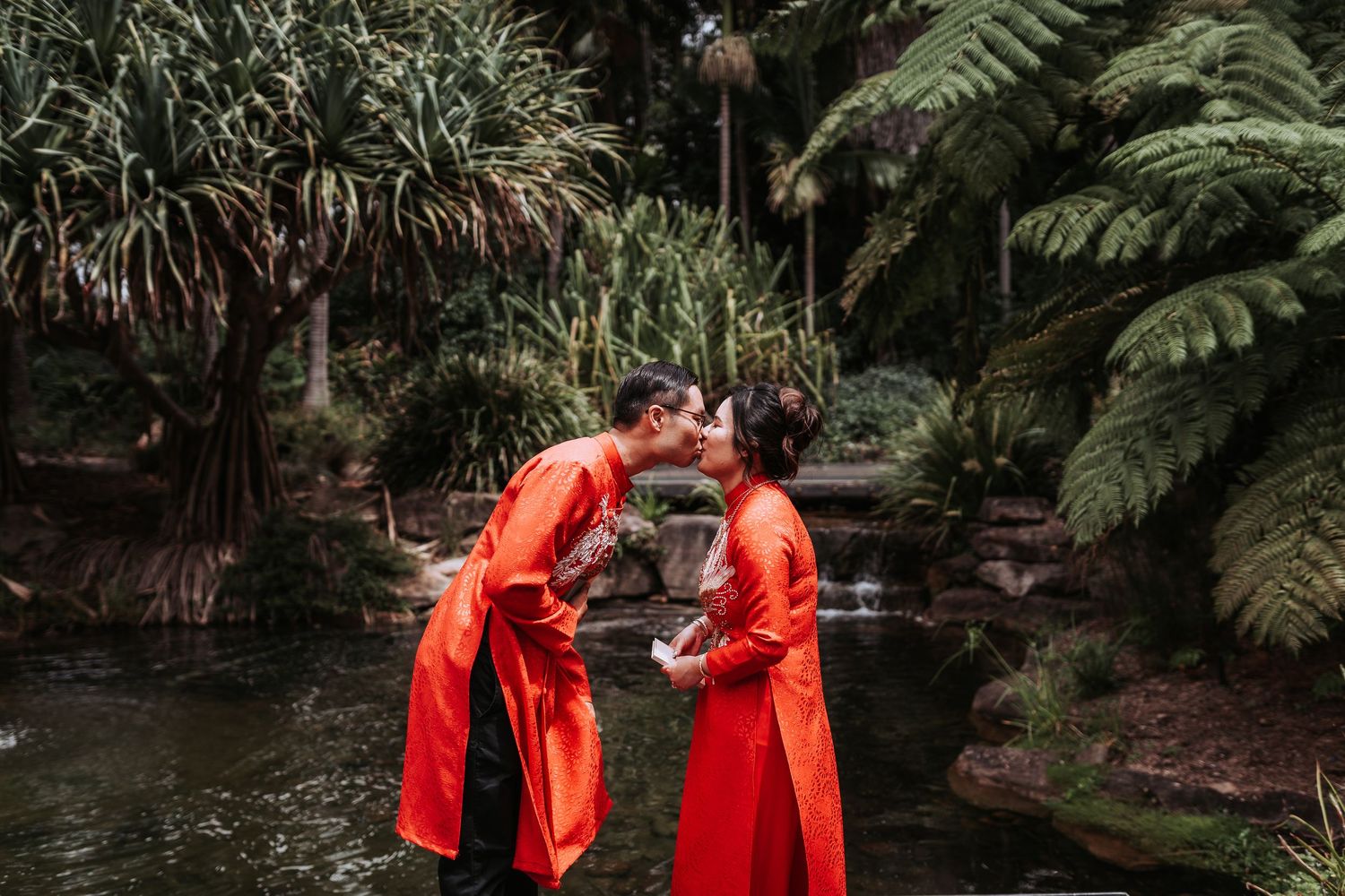 A couple in matching red traditional outfits stands together by a pond surrounded by lush ferns.