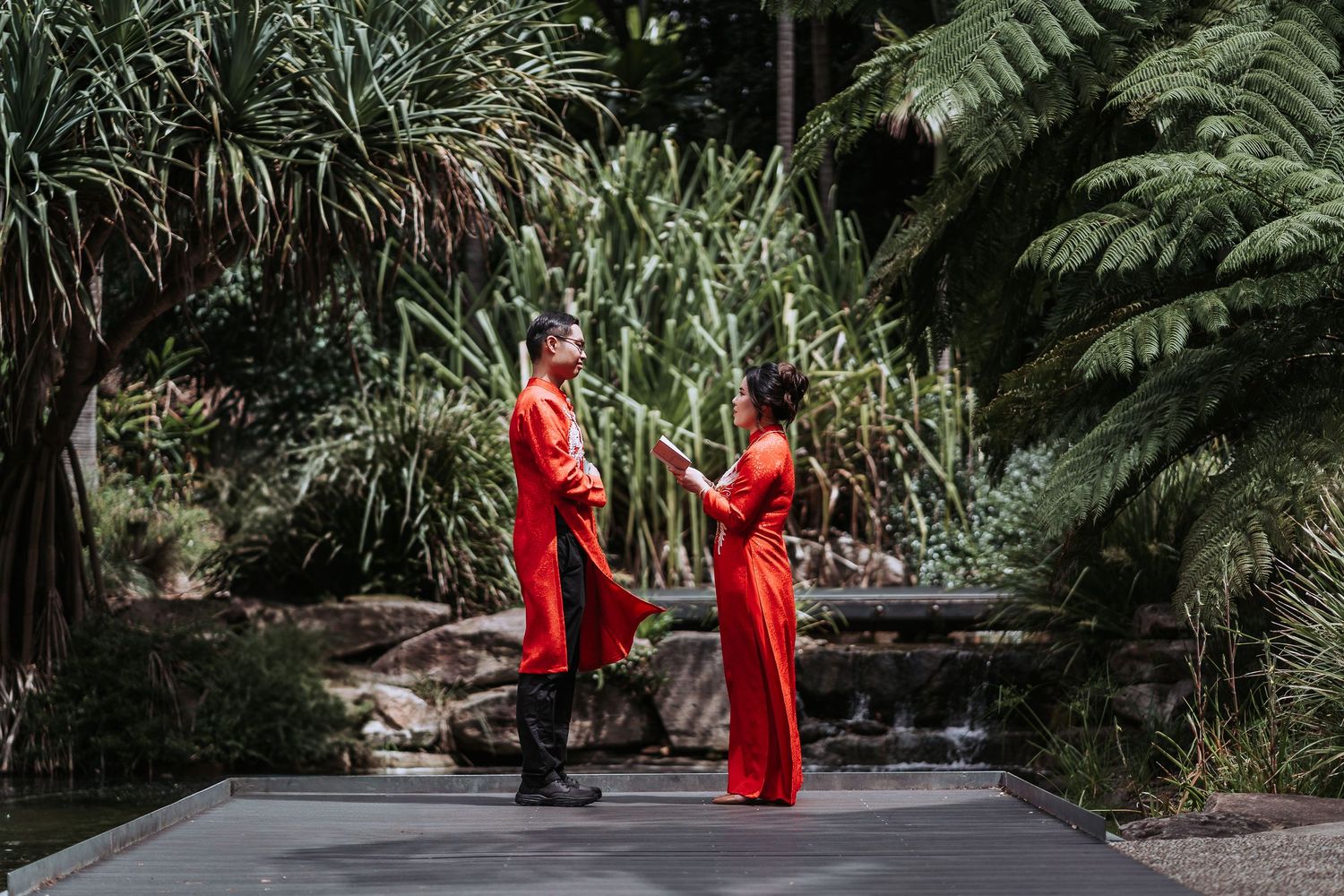 Couple in matching red attire standing on wooden boardwalk surrounded by lush tropical foliage.