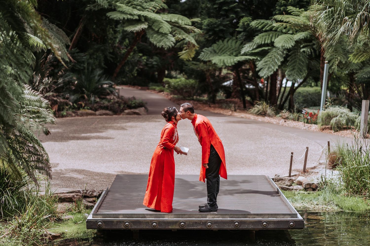 Two people in bright red traditional clothing share an intimate moment on wooden deck in garden setting.