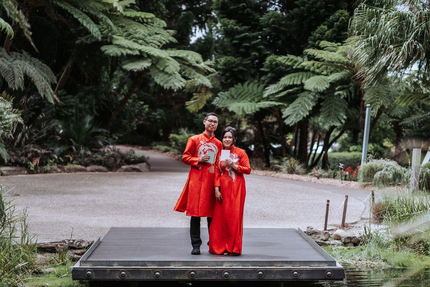 Couple in matching red traditional outfits walking together on a wooden pathway surrounded by lush greenery.
