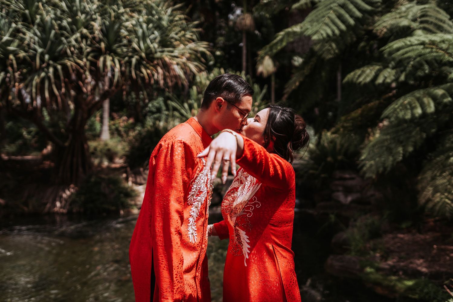 A couple in matching bright red traditional Vietnamese wedding outfits embrace near a pond in a lush garden.