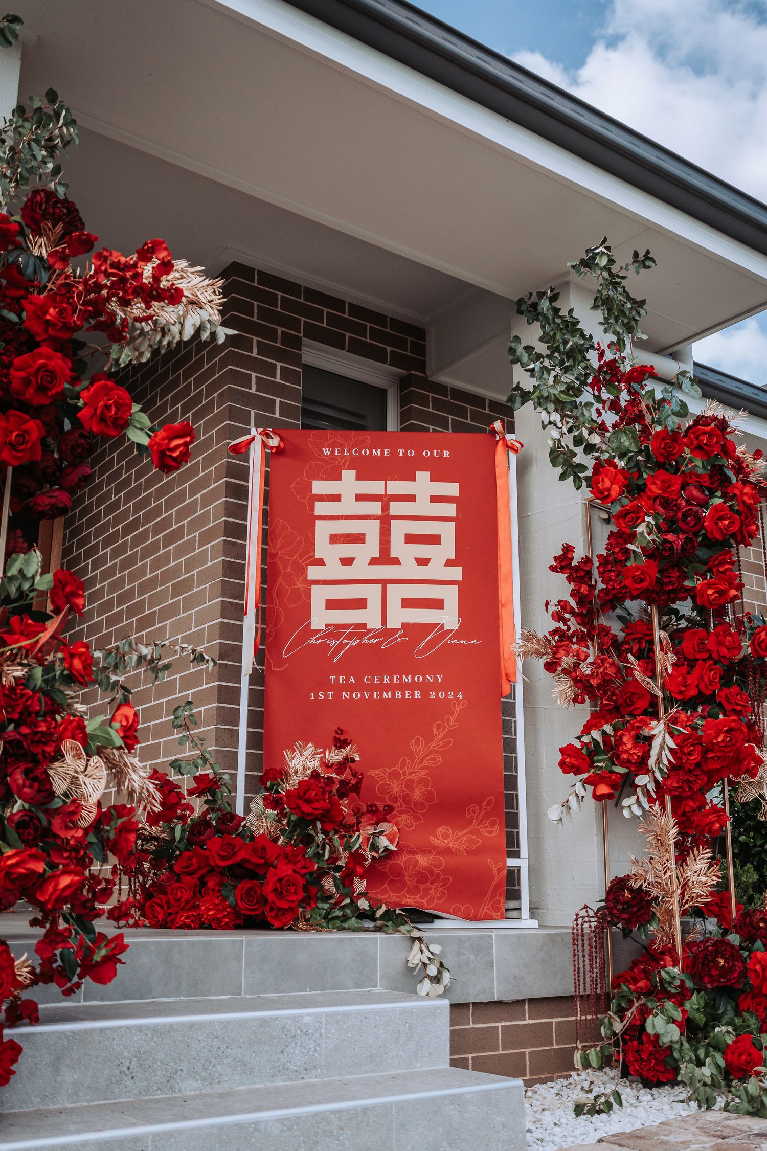 Red wedding banner with double happiness symbol displayed on brick exterior surrounded by vibrant red climbing roses.