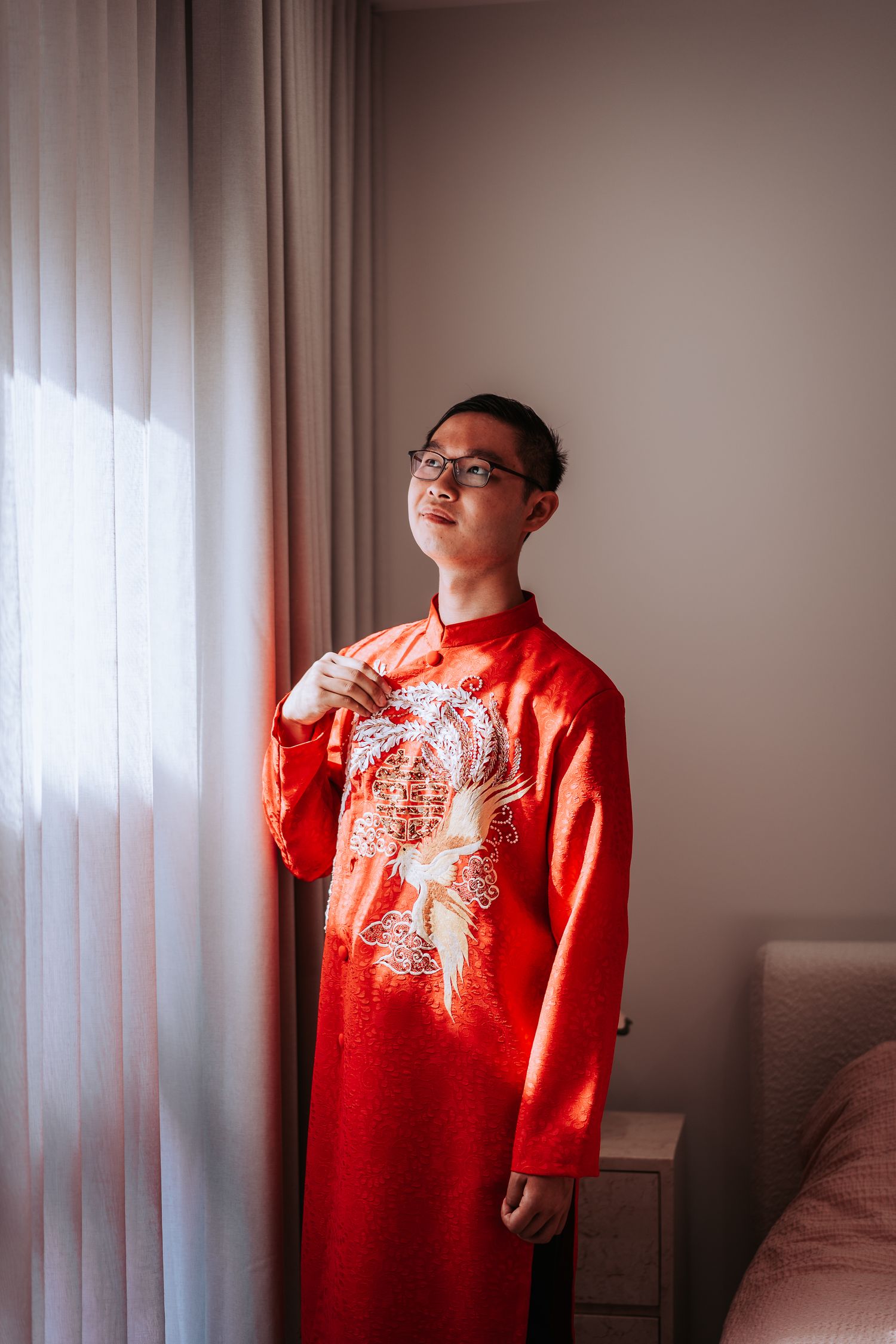 Person wearing traditional red Chinese New Year attire with dragon embroidery stands near window in natural lighting.
