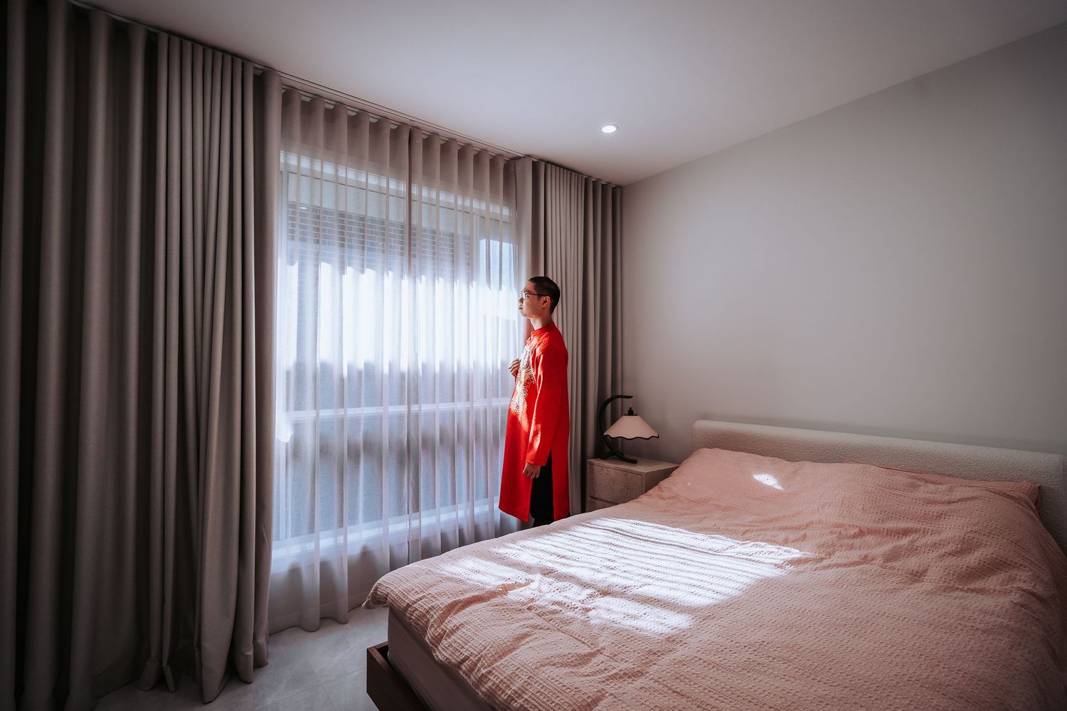 A person in a red dress stands by a window in a modern bedroom with grey curtains.