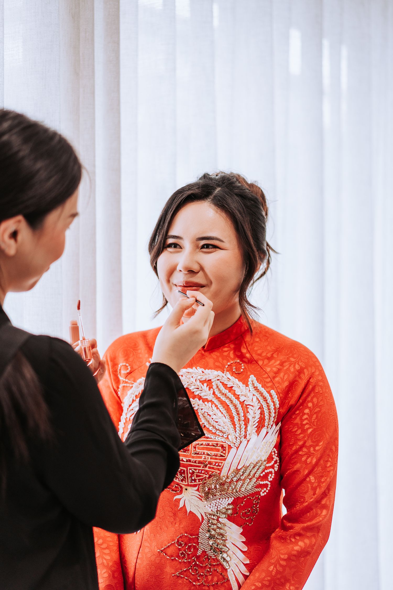 A person in a vibrant red traditional Chinese dress with crane embroidery gets their makeup done against white curtains.