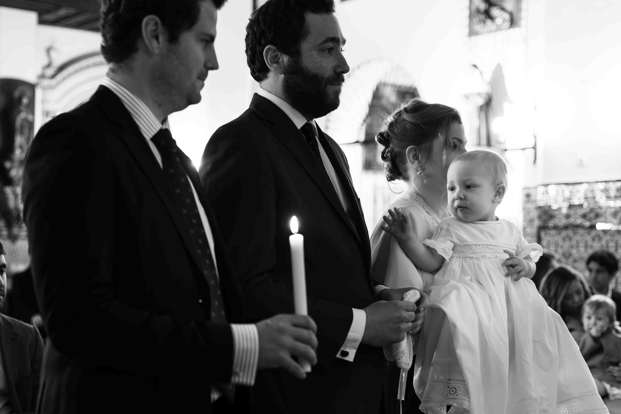 A black and white photograph of a baptism ceremony showing adults holding candles beside a child in white christening gown.