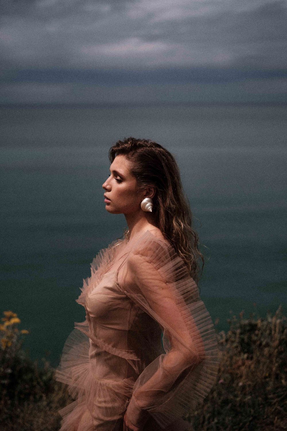 An ethereal portrait in a blush tulle dress against a moody coastal landscape.