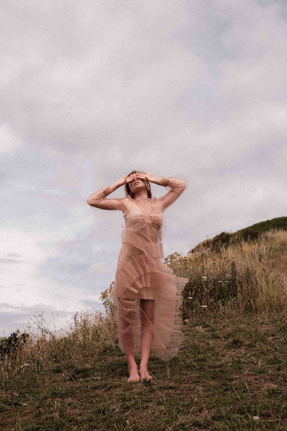 Figure in sheer blush dress poses gracefully on hillside under cloudy sky.