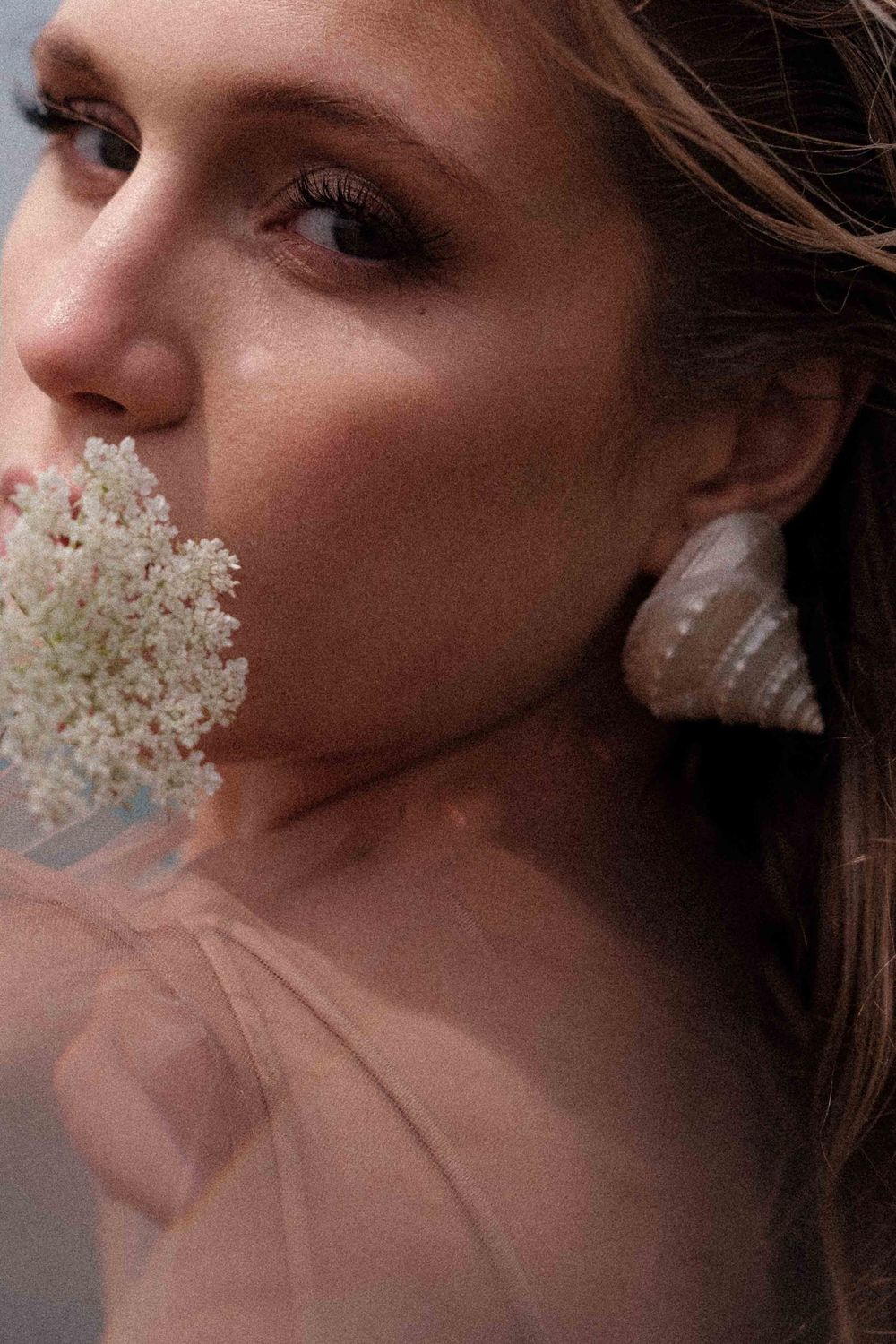 Artistic portrait with white flowers and seashell earring against blush tones.