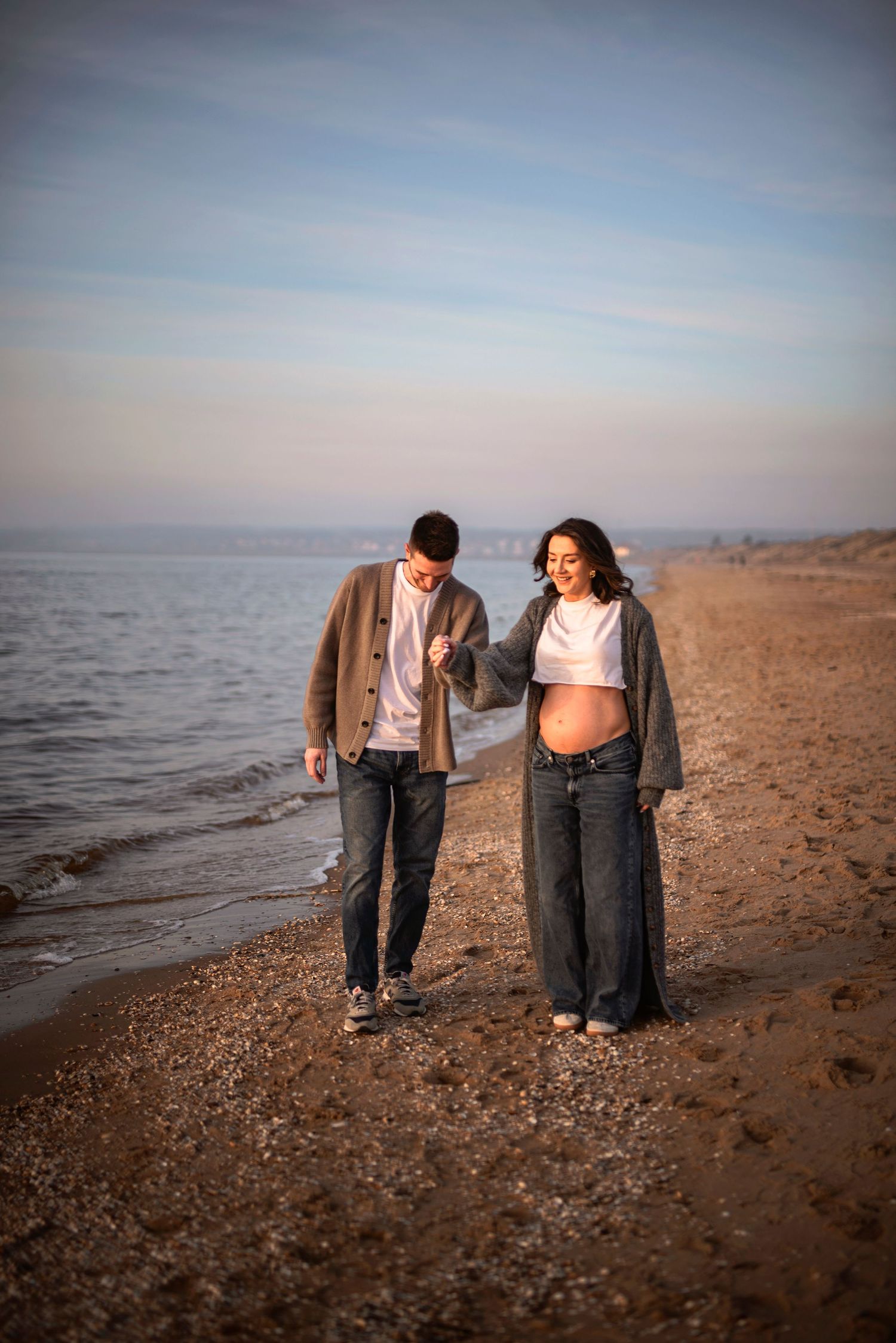 Two people walk together on a beach shoreline at sunset wearing casual clothing and sweaters.