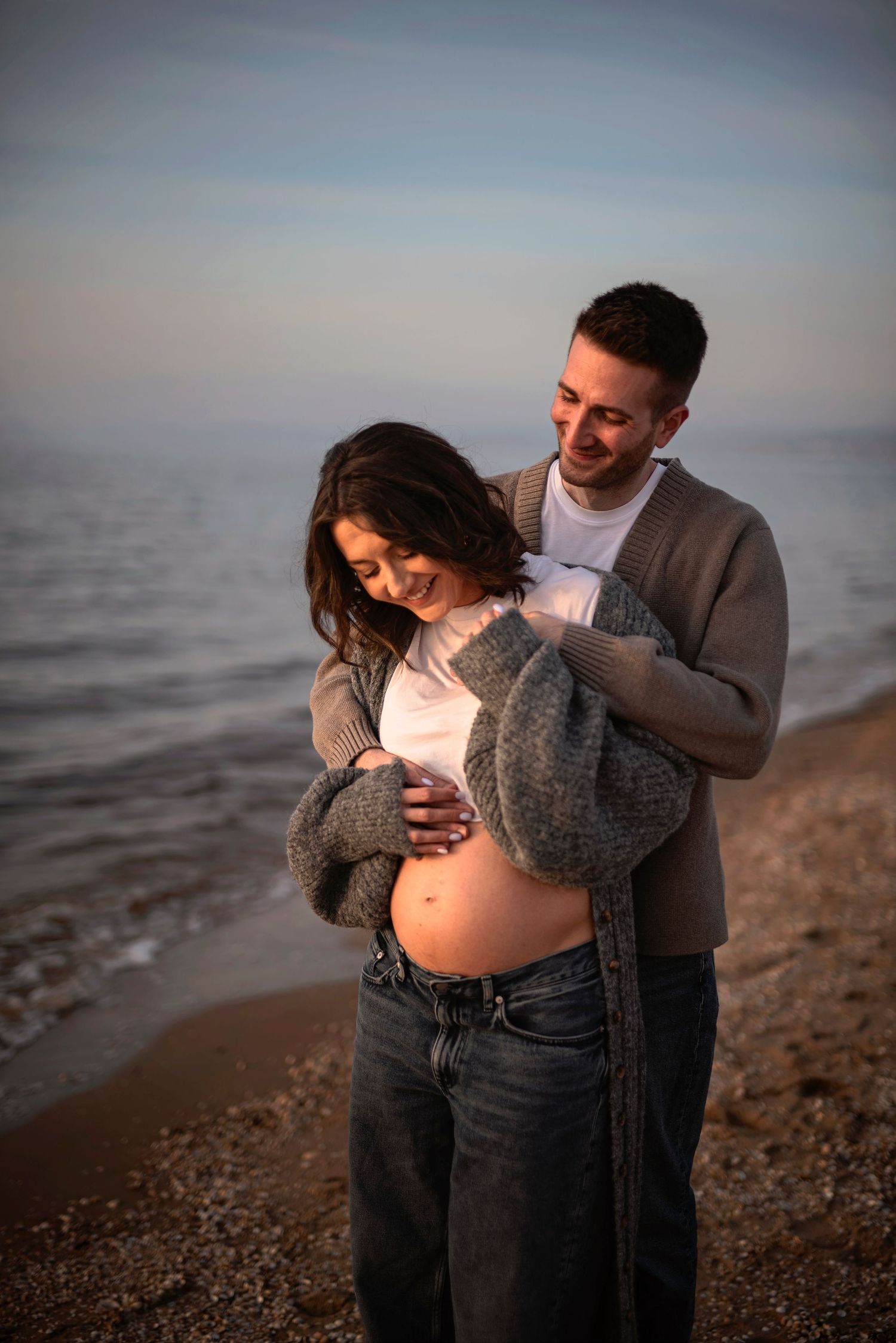 Expecting couple shares intimate moment on beach at sunset as they embrace her pregnant belly.