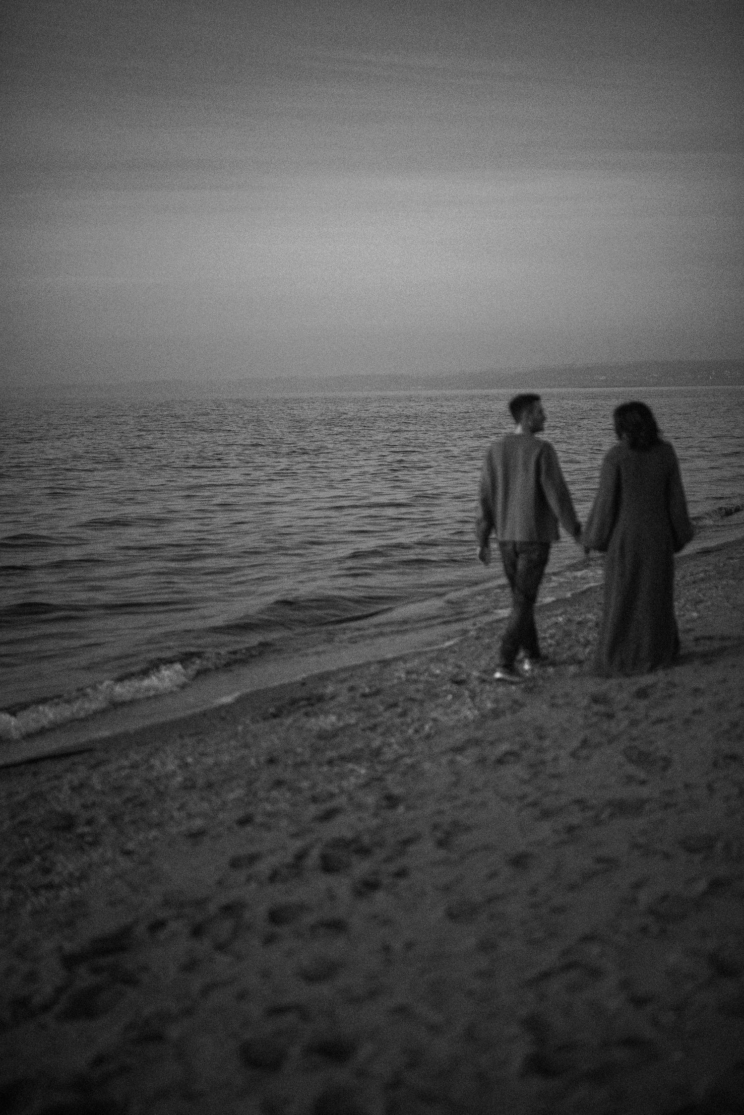 A couple walks hand in hand along a beach shoreline at dusk in black and white.