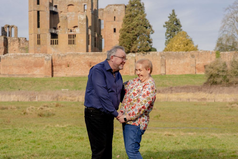 Two people share a tender moment while standing on grass in front of an ancient stone castle on an autumn day.