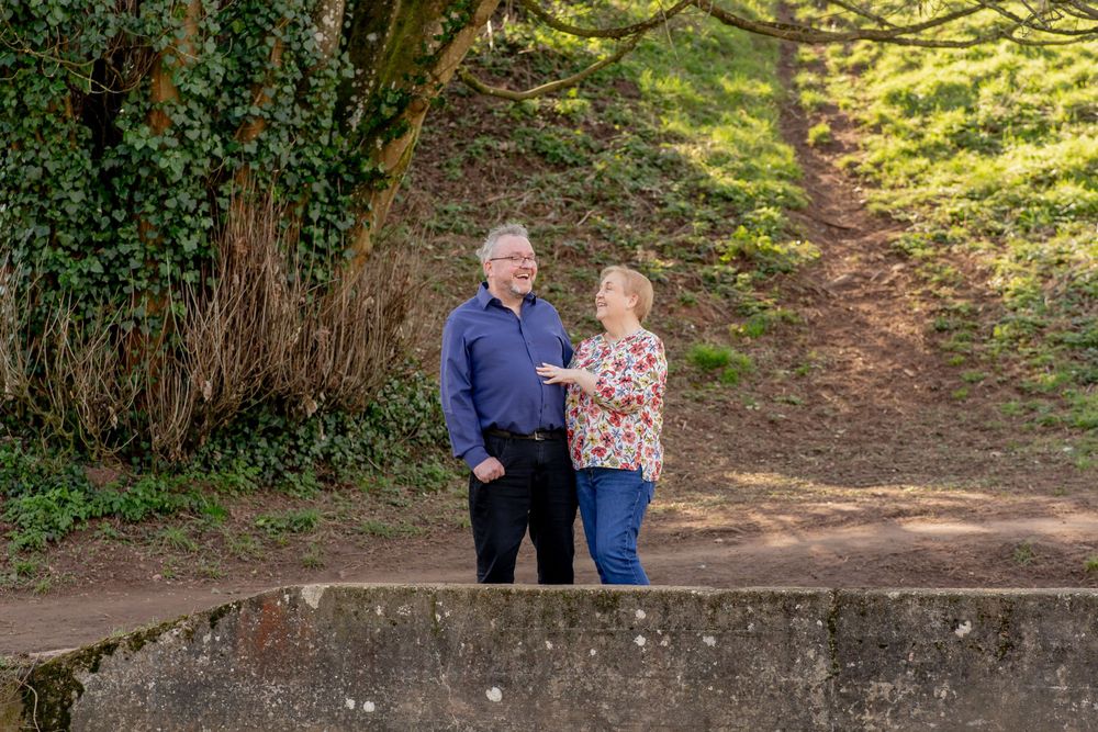 A couple stands together on a sunny woodland path with ivy-covered trees and dappled sunlight in the background.