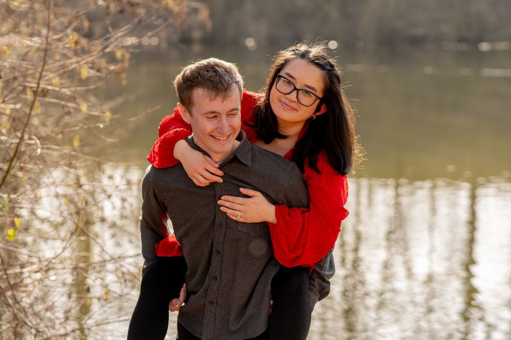 A couple shares a playful piggyback moment by a serene lake on an autumn day.