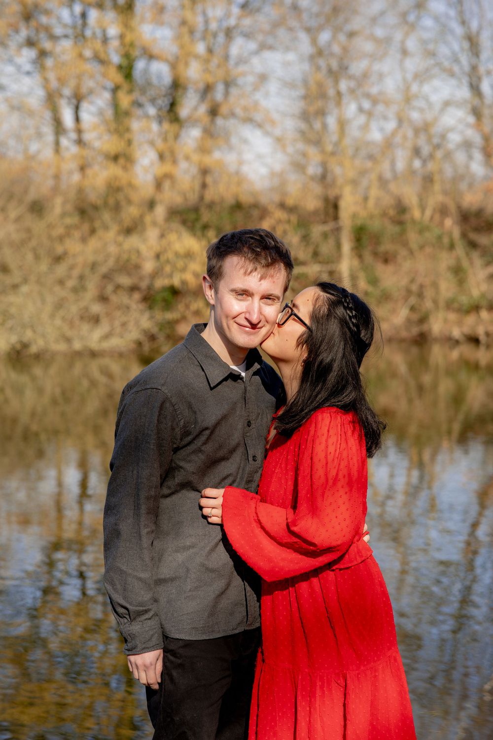 A couple shares a romantic moment by a lake on an autumn day, with the person in red embracing their partner.