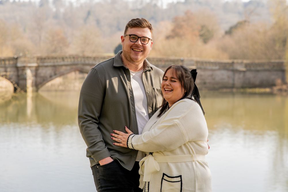 A happy couple embraces while standing by a lake with a stone bridge in the background on an autumn day.