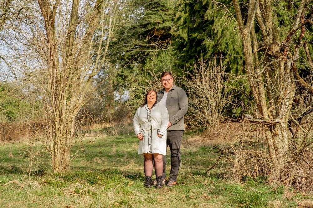 A couple stands together outdoors on a grassy hillside surrounded by bare winter trees in warm afternoon light.