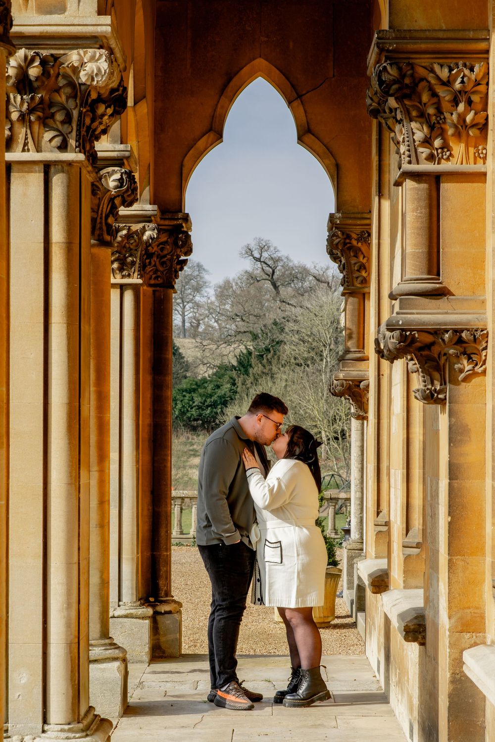 Romantic couple embracing beneath a Gothic-style archway with decorative stone columns and capitals.