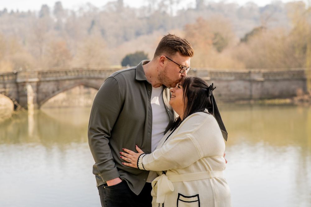 A romantic winter portrait of a couple embracing by a serene lake with a stone bridge in the background.