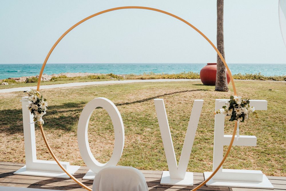 Modern circular wedding arch with white LOVE letters sits against ocean backdrop on grassy venue.