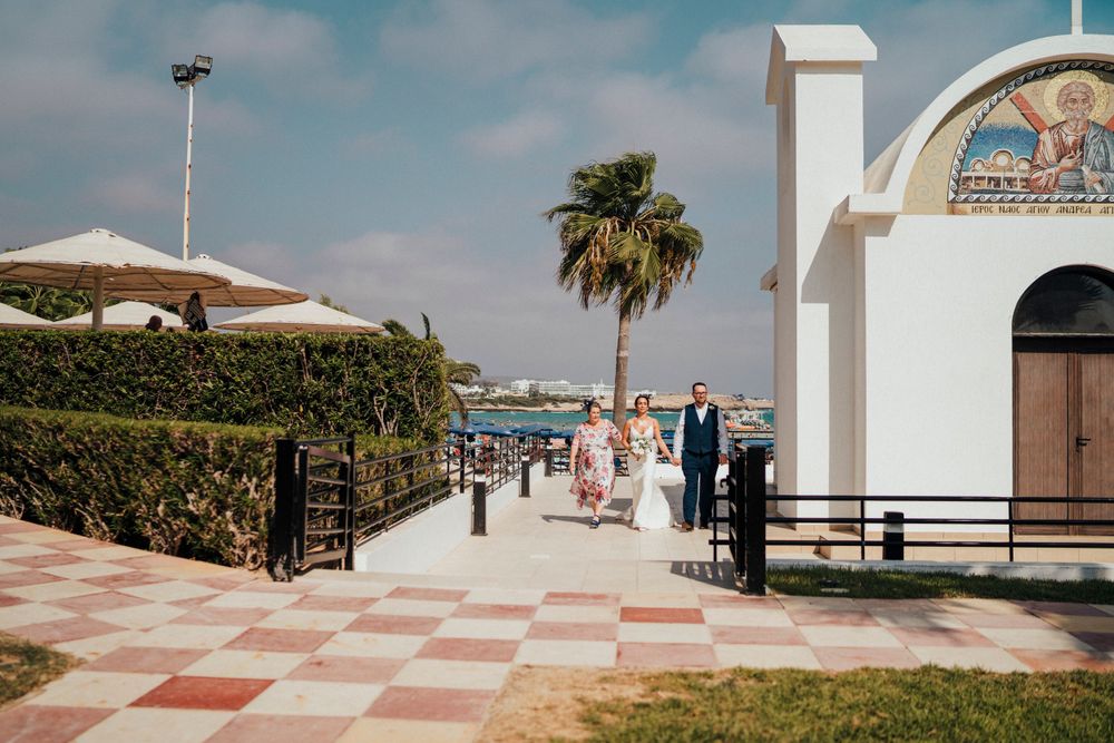 Outdoor beach promenade with palm tree and white arched building entrance near umbrellas and hedges on sunny day.