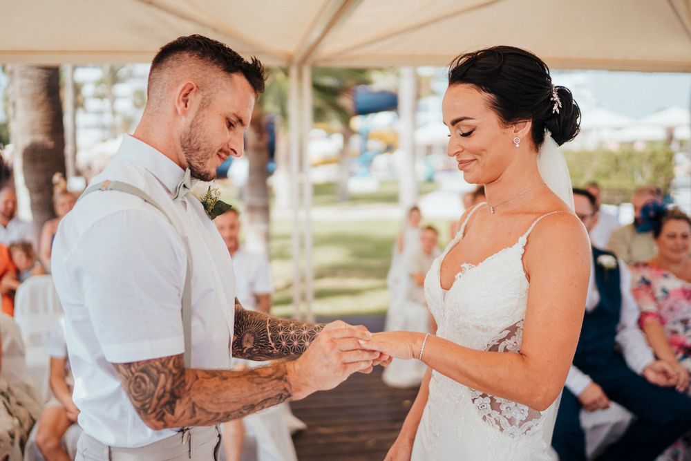 A couple exchanges rings during a beach wedding ceremony under a white canopy tent.