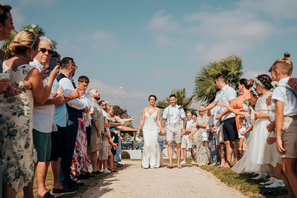 Wedding guests line a walkway outdoors as they blow bubbles at a sunny beach celebration ceremony.
