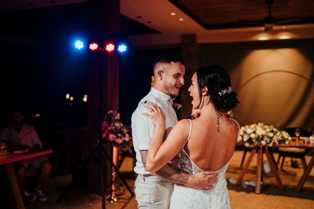 Newlyweds share their first dance together at a wedding reception with blue stage lights and floral decor in background.