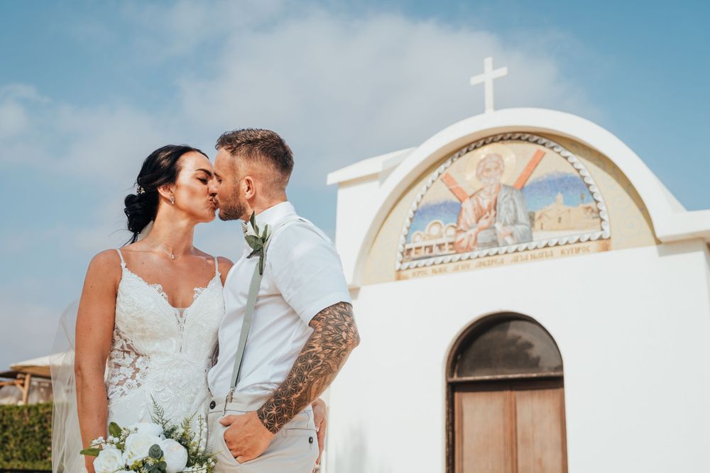 A couple in wedding attire share an intimate moment in front of a white Mediterranean church with a cross on top.