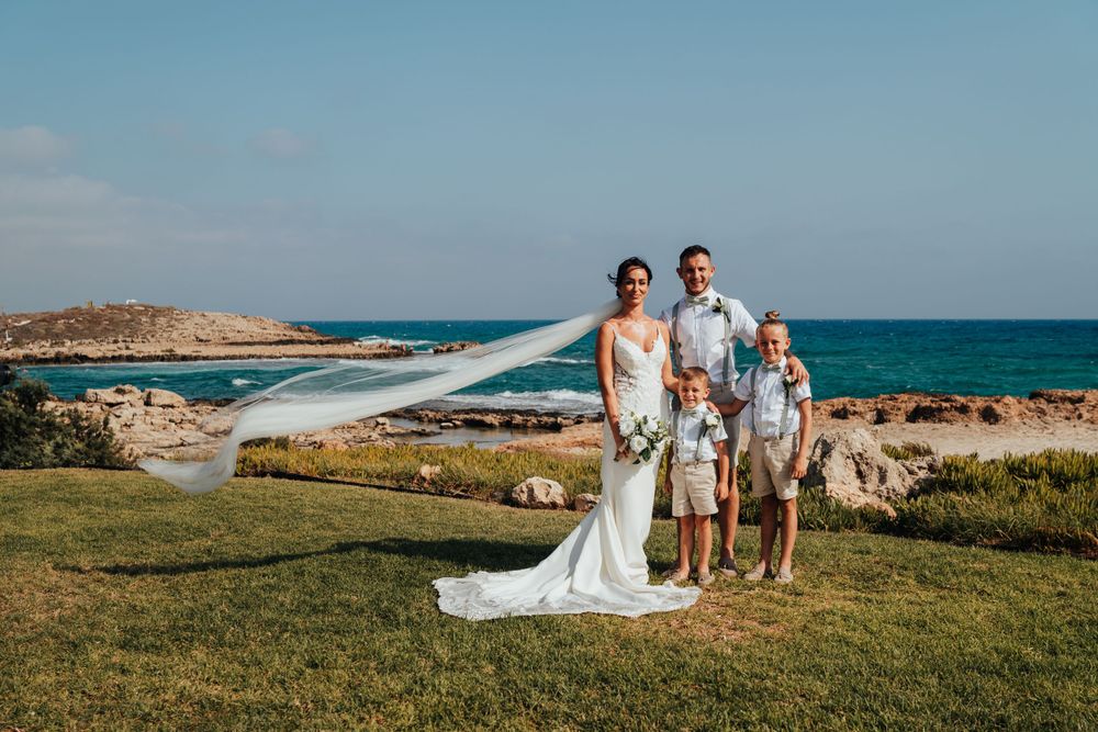 Wedding group stands on scenic coastal cliff with ocean and blue sky backdrop, bride's veil flowing in wind.