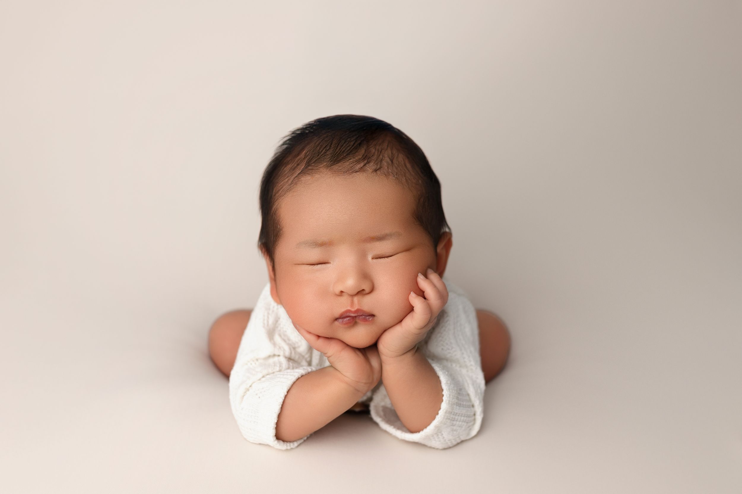 Sleeping newborn baby in white outfit lying on stomach with hands propped under chin on cream backdrop.