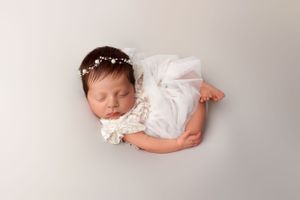 A sleeping newborn wearing a delicate white lace dress and small pearl headband rests peacefully on a light gray backdrop.