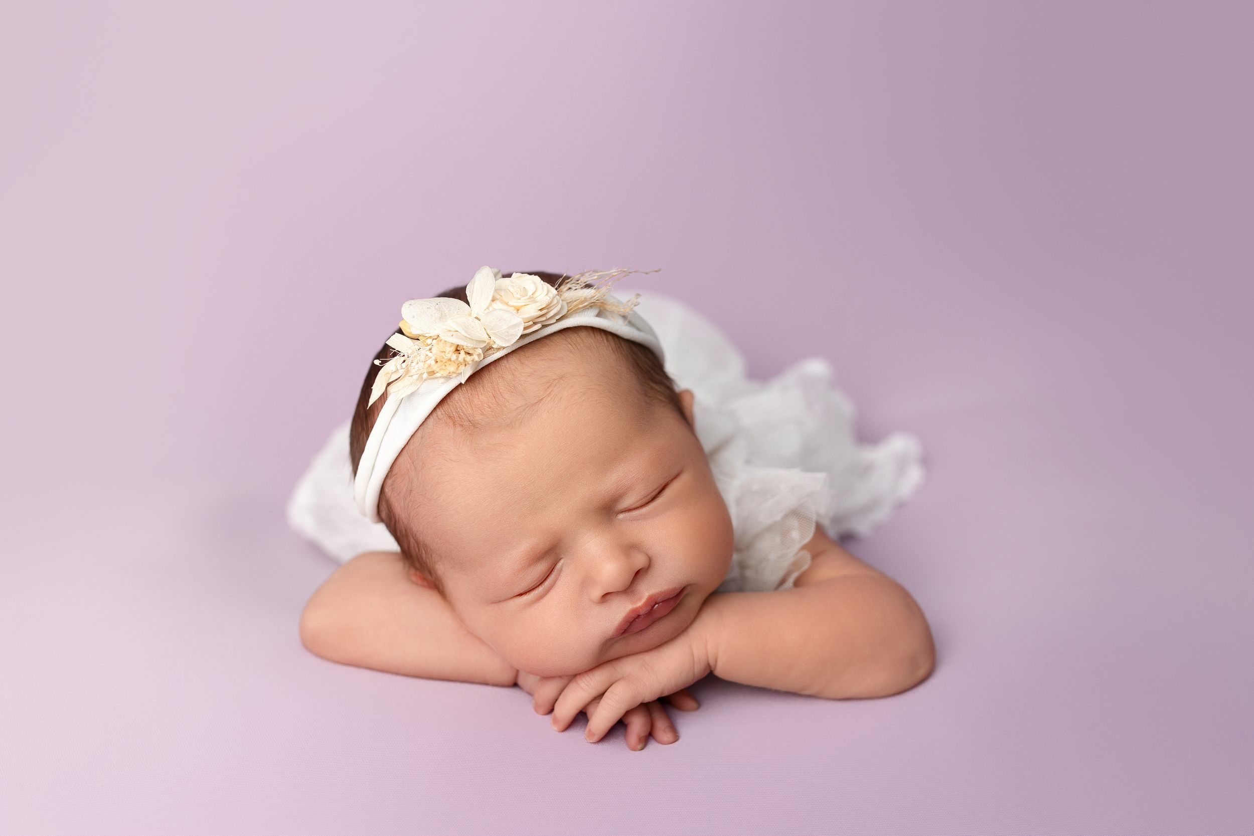 Peaceful sleeping newborn wearing white headband rests on soft pink background.