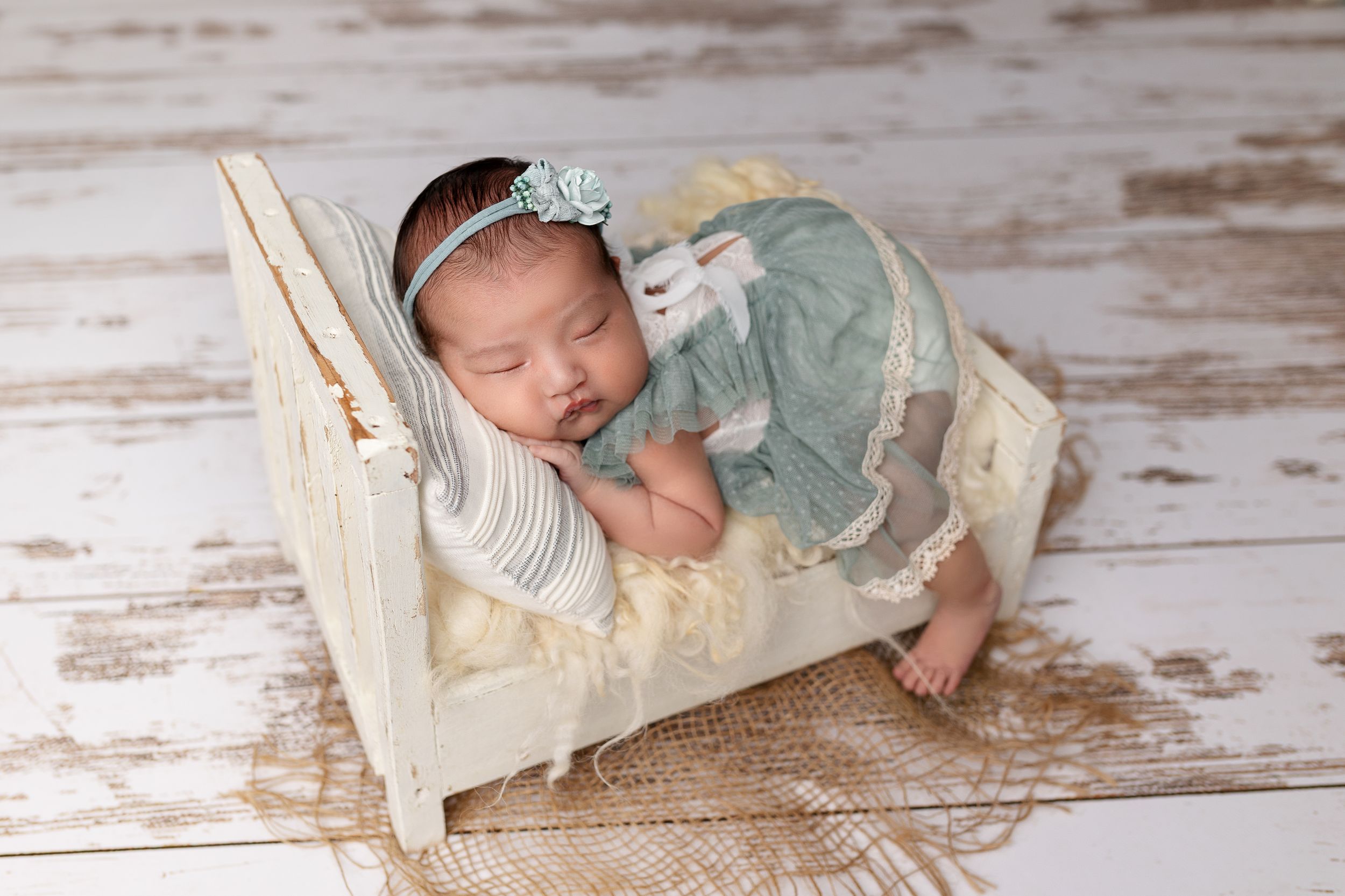 A sleeping newborn wearing a silver headband and mint green outfit rests peacefully in a vintage white wooden bed.