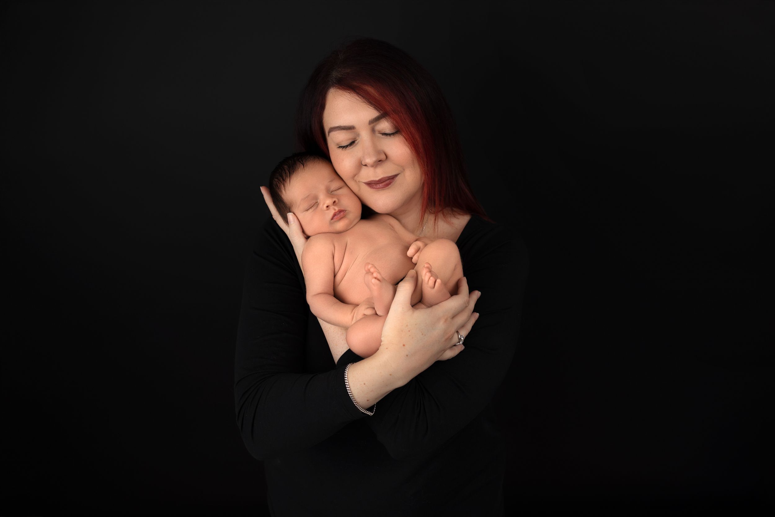 A tender moment captured against a dark background as a parent in black clothing cradles their sleeping newborn baby.