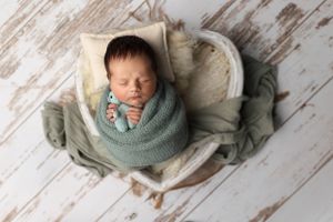 Sleeping newborn wrapped snugly in a sage green swaddle on rustic white wooden backdrop.