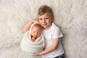 A young child in a white shirt poses with a newborn swaddled in a white wrap on a white textured background.