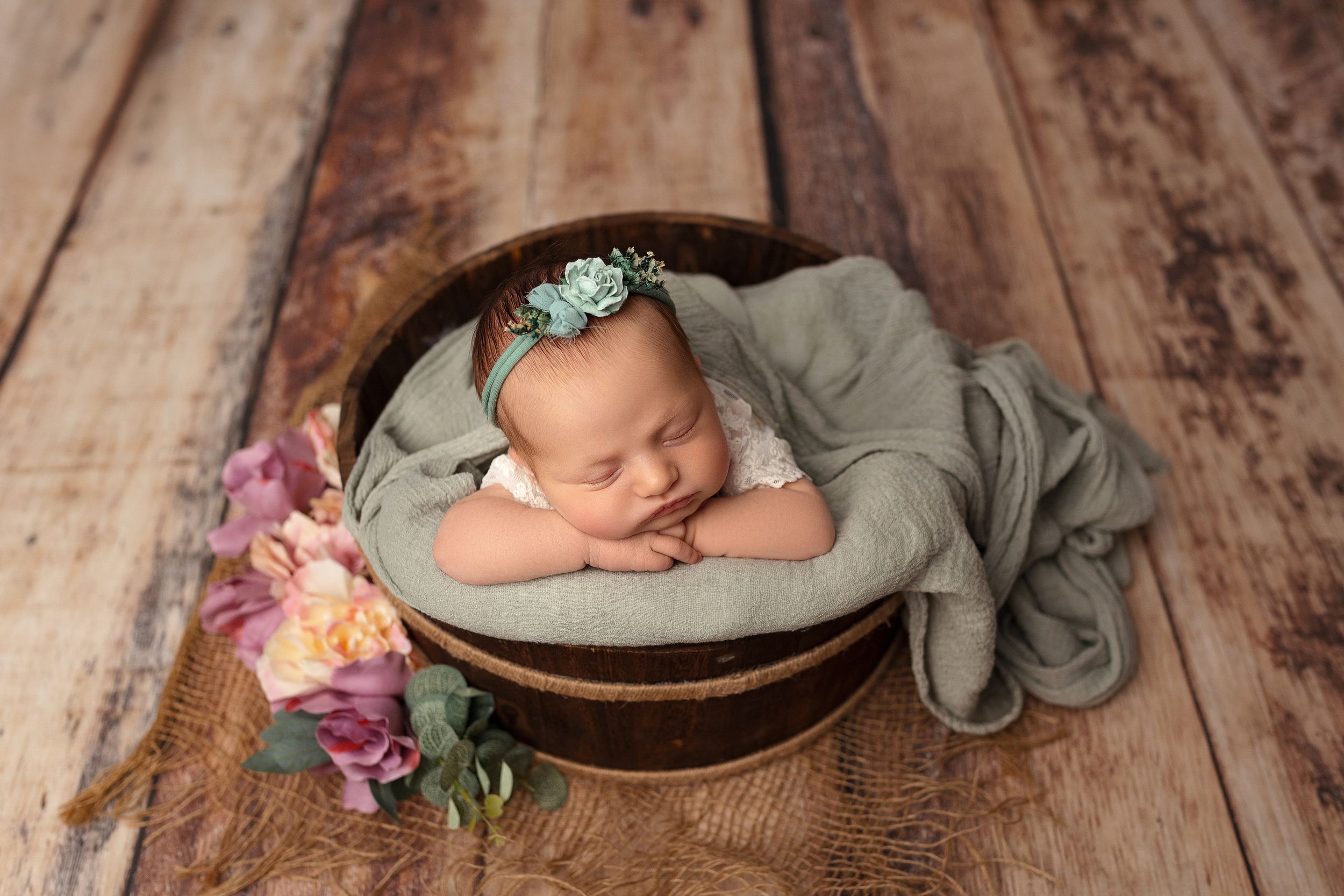 Sleeping newborn in sage green wrap posed in round basket with floral decor on wooden floor backdrop.