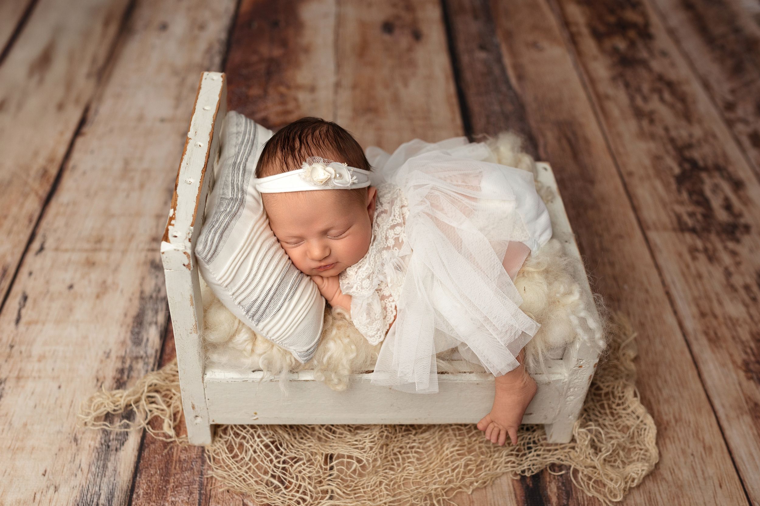 A newborn sleeps peacefully in a rustic white wooden bed with lace dress and headband on wooden floor backdrop.