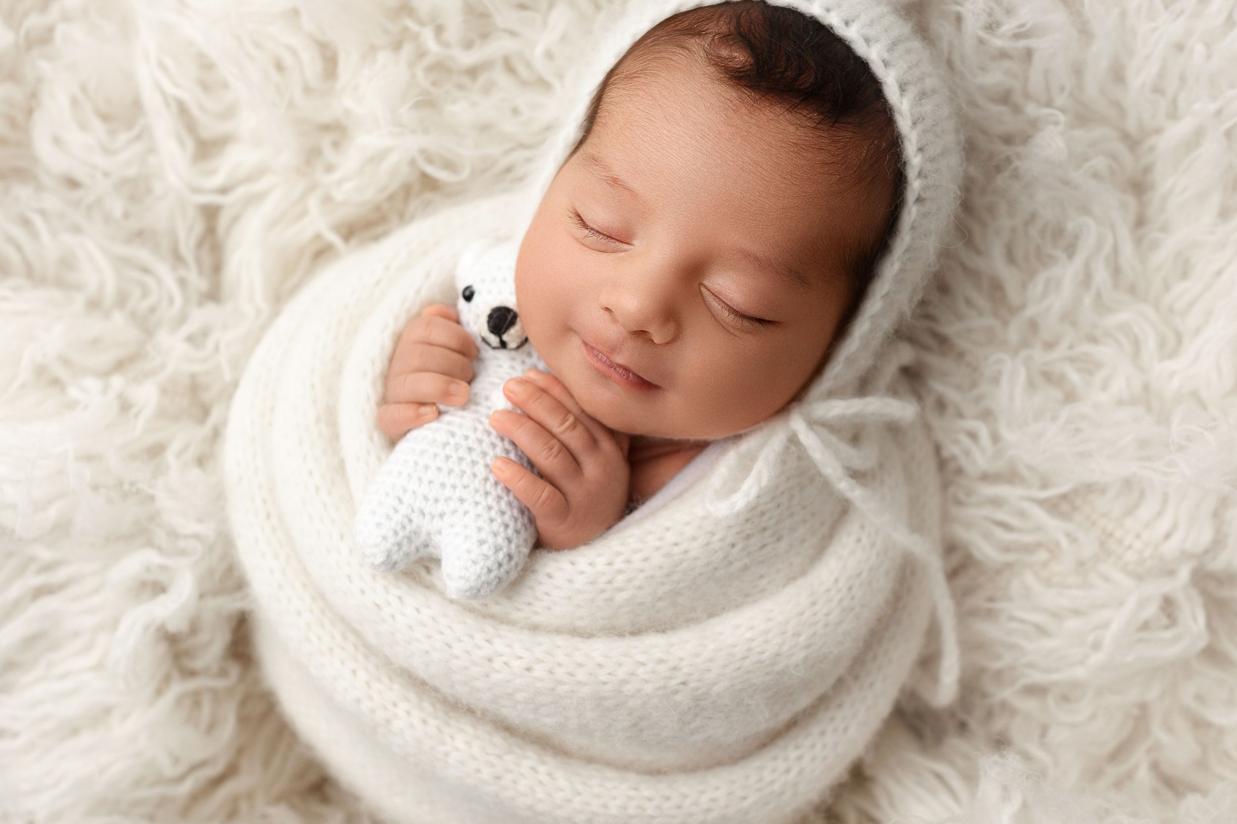 A peaceful sleeping newborn wrapped in soft white blanket rests on plush fur background.